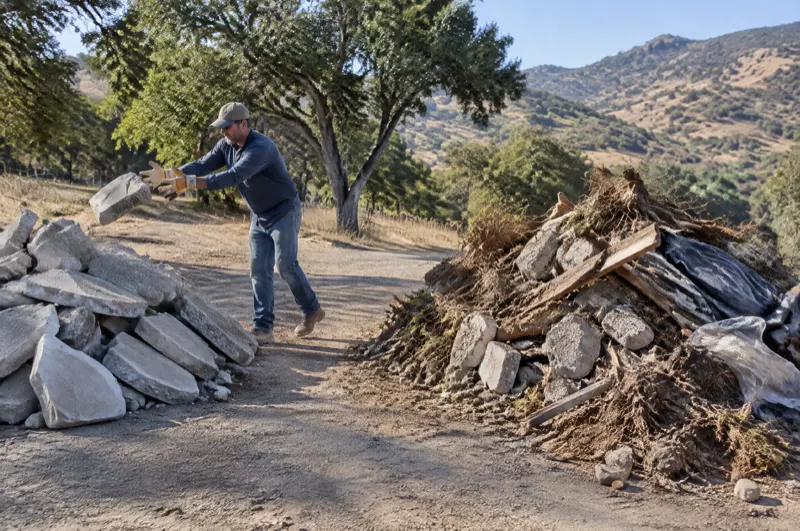 Worker tossing a slab piece onto a clean concrete pile beside a mixed pile of concrete, roots, wood and plastic